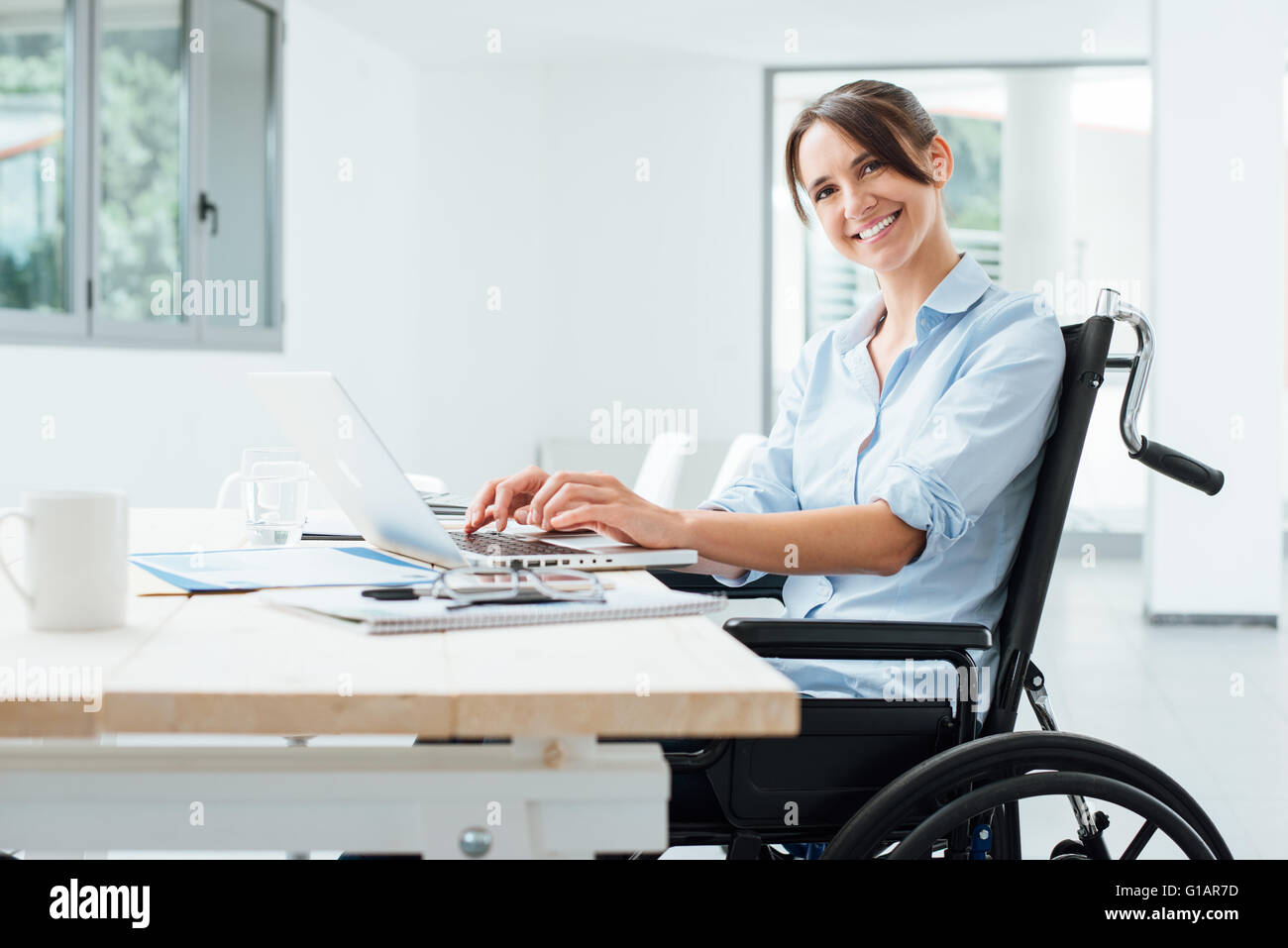 Confident happy woman in wheelchair travaillant au bureau et à l'aide d'un ordinateur portable, il est smiling at camera, un handicap plus Banque D'Images