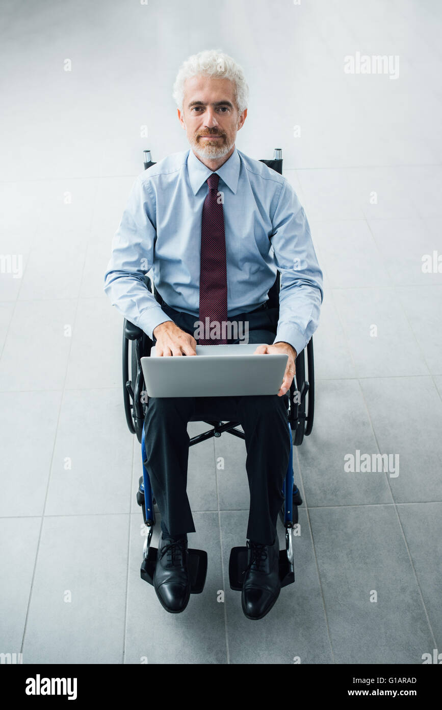 Confiant smiling businessman en fauteuil roulant à l'aide d'un ordinateur portable, l'arrière-plan sur l'intérieur de bureau Banque D'Images