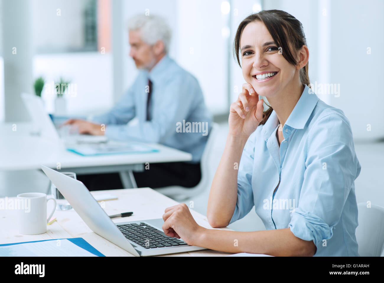 Smiling young business woman sitting at Office 24 et travailler avec un ordinateur portable, elle est looking at camera Banque D'Images