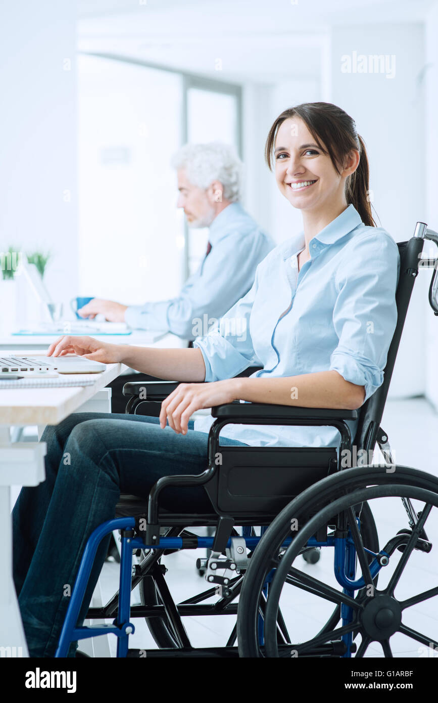Confident happy woman in wheelchair travaillant au bureau et à l'aide d'un ordinateur portable, il est smiling at camera, un handicap plus Banque D'Images