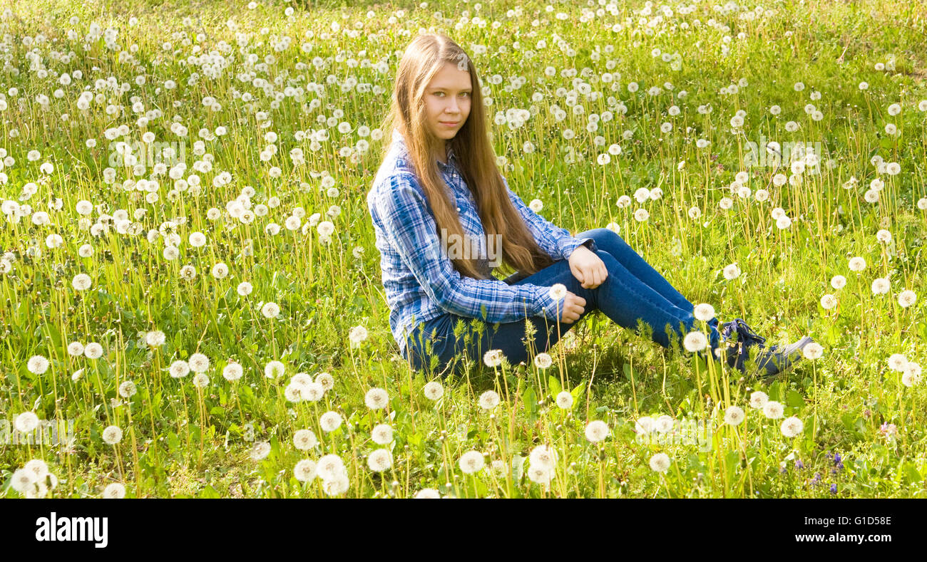 Belle jeune fille, seize ans, Européen, avec de longs cheveux bruns, en jeans et jupe, siège au pré avec pissenlits blancs. Banque D'Images