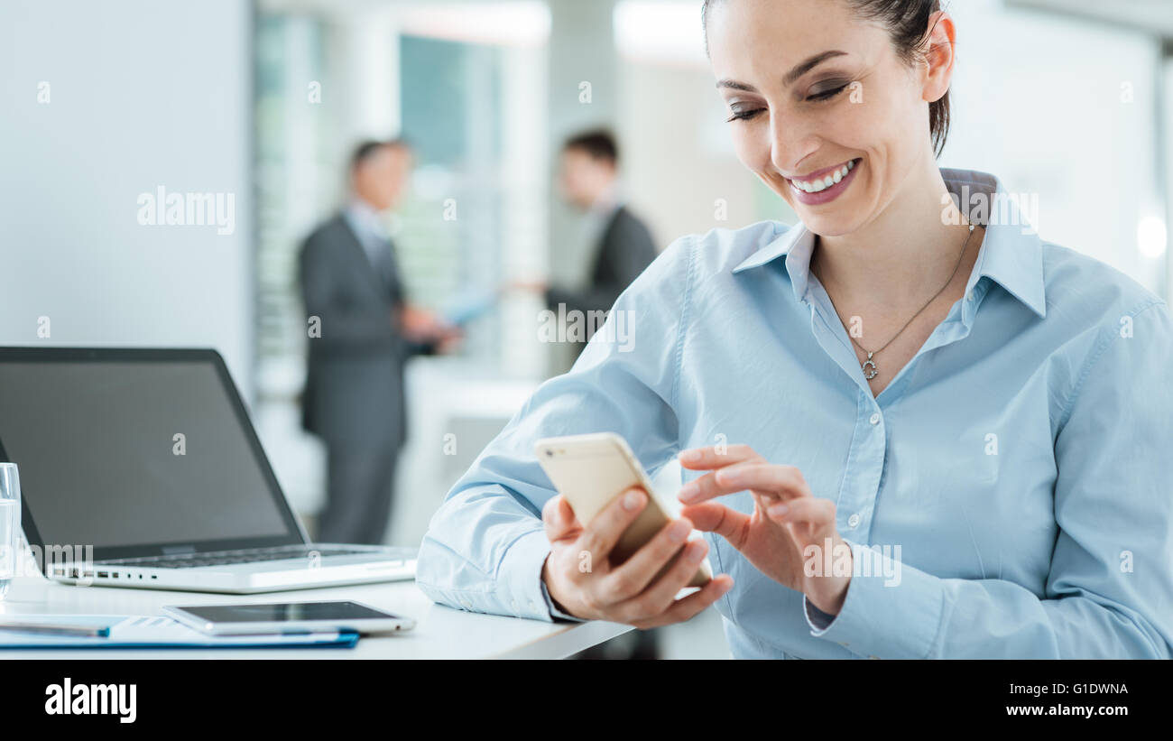 Confiant smiling business woman sitting at Office 24 et à l'aide d'un téléphone intelligent, des gens d'affaires debout sur arrière-plan, Banque D'Images