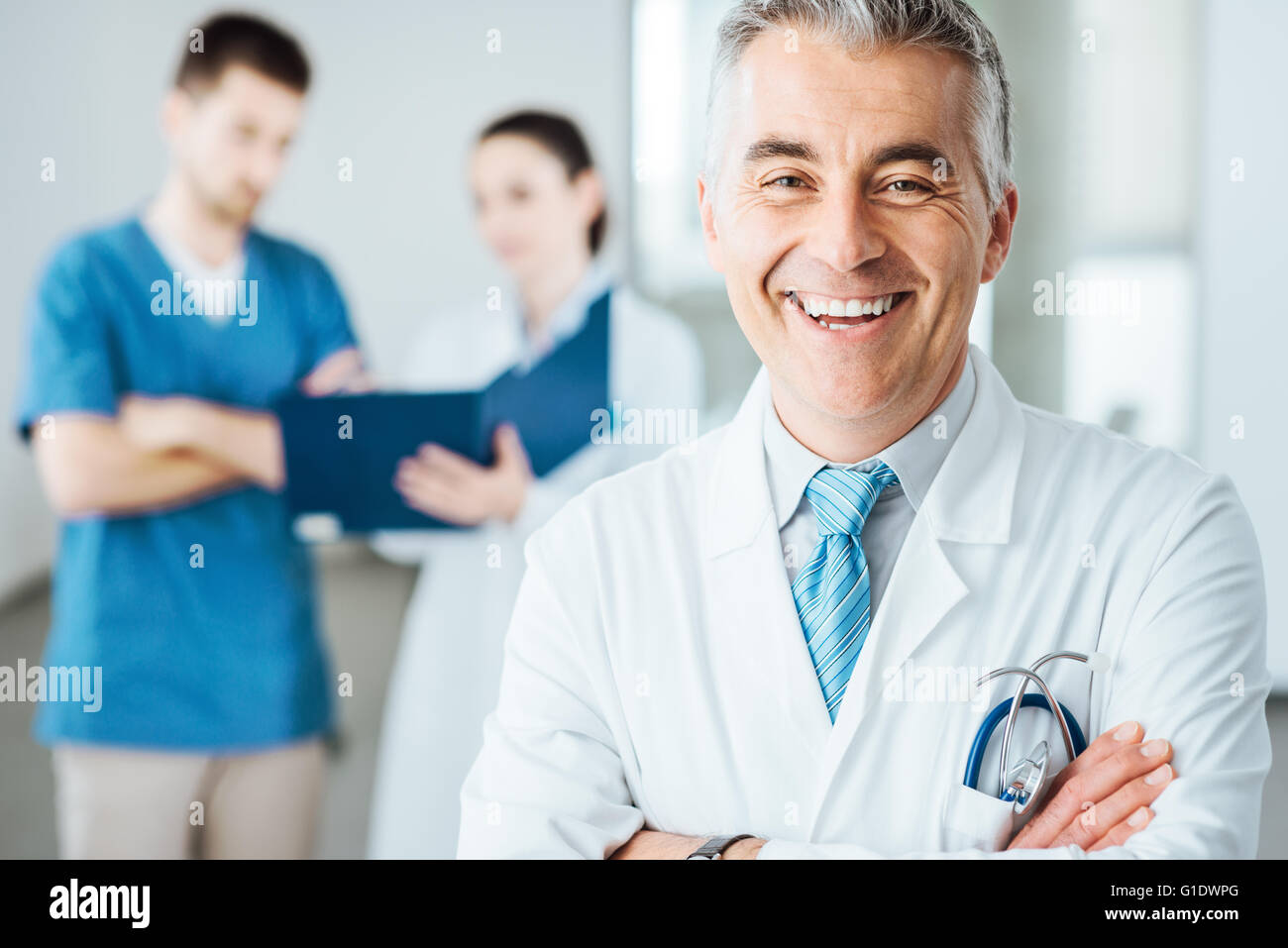 Confiant doctor posing and smiling at camera et le personnel médical contrôle de dossiers médicaux sur l'arrière-plan Banque D'Images