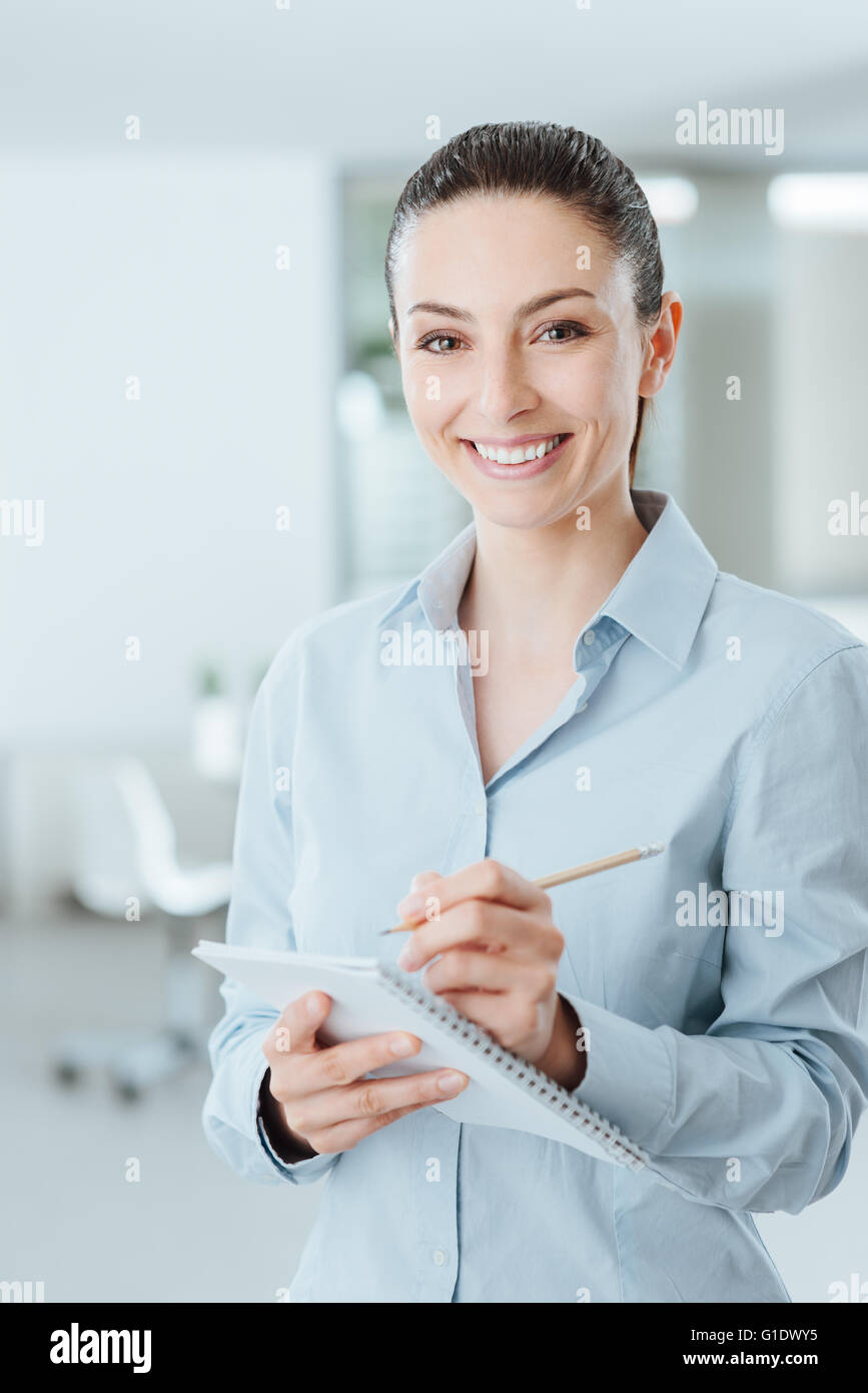 Jeune secrétaire professionnel écrit des notes sur un ordinateur portable et smiling at camera, elle se tient dans le bureau Banque D'Images