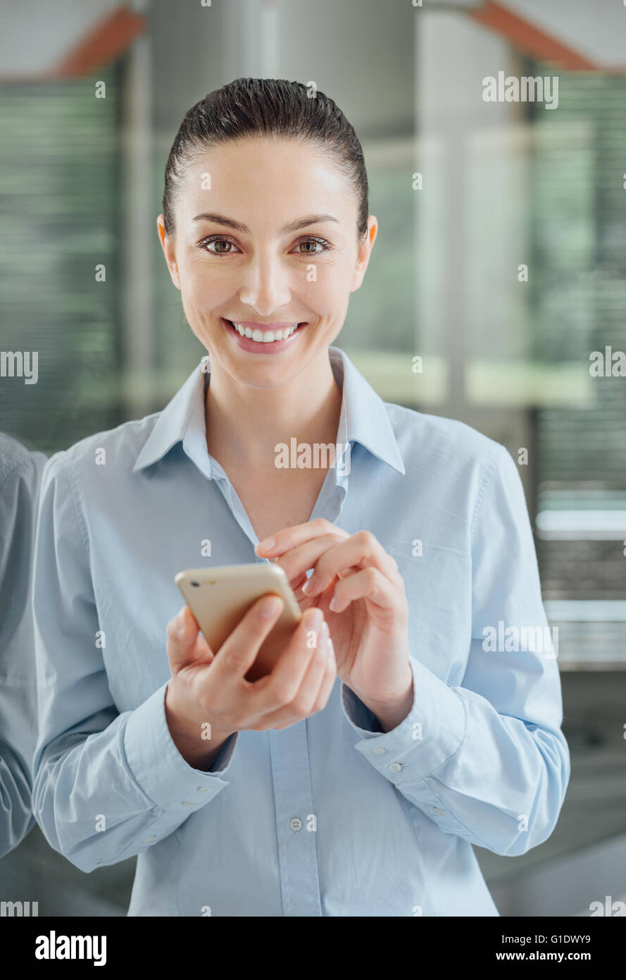 Belle jeune femme à l'aide d'un téléphone intelligent et s'appuyant sur une fenêtre, elle est smiling at camera Banque D'Images