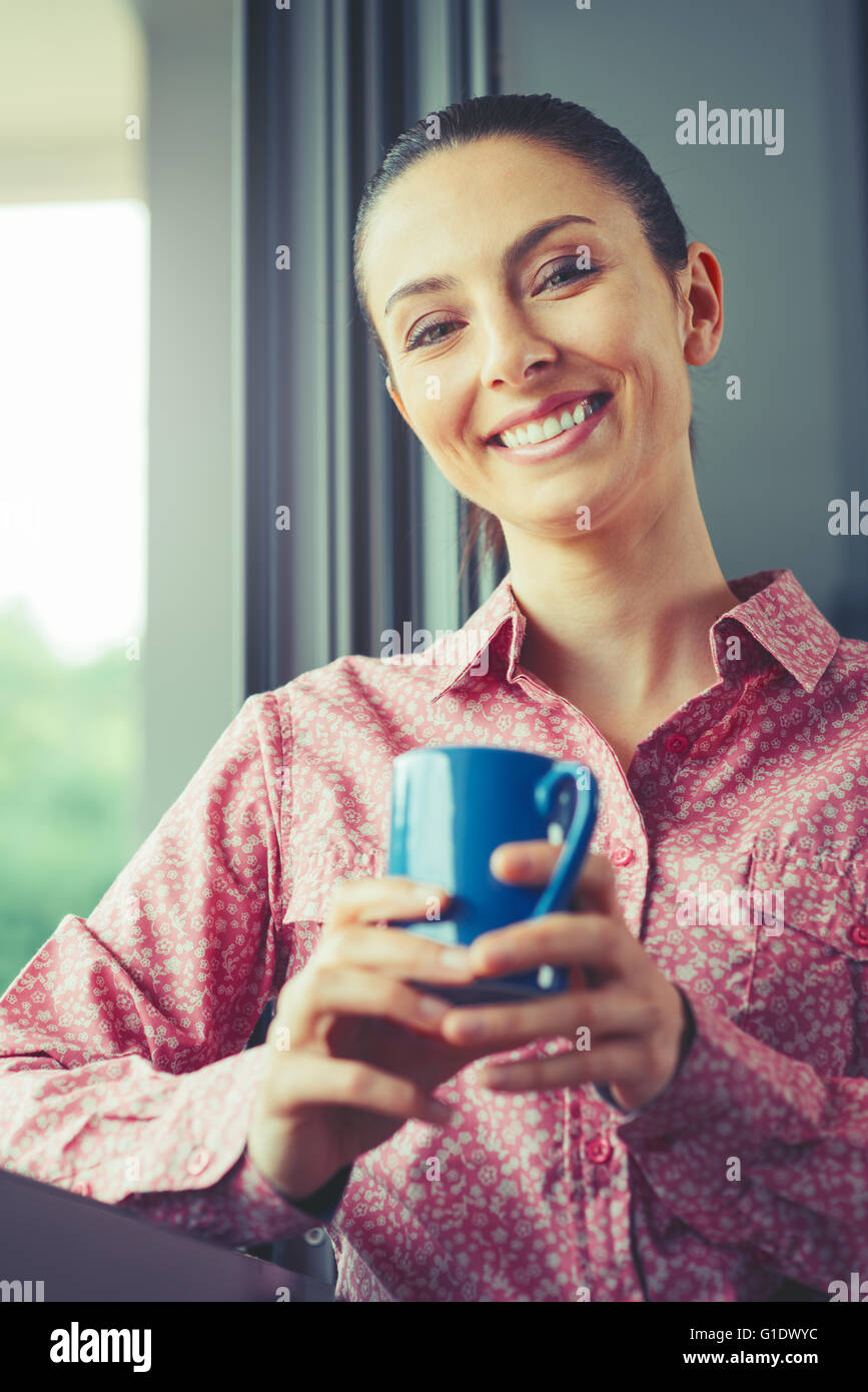 Femme ayant une pause café à fenêtre, elle est souriante et tenant une chope Banque D'Images