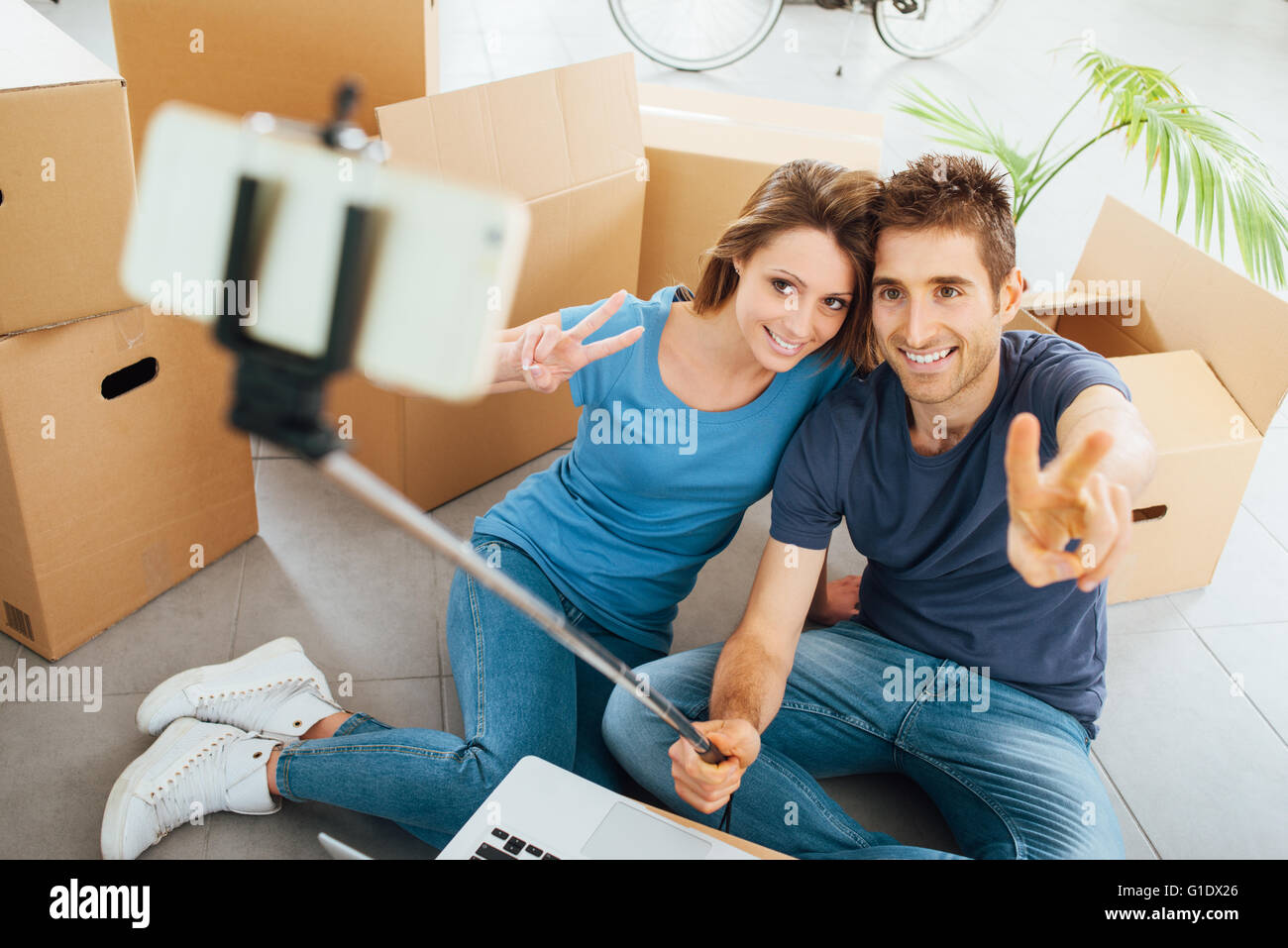 Smiling young couple sitting on leur nouvelle maison étage entouré de cartons et prendre des autoportraits à l'aide d'un bâton selfies Banque D'Images