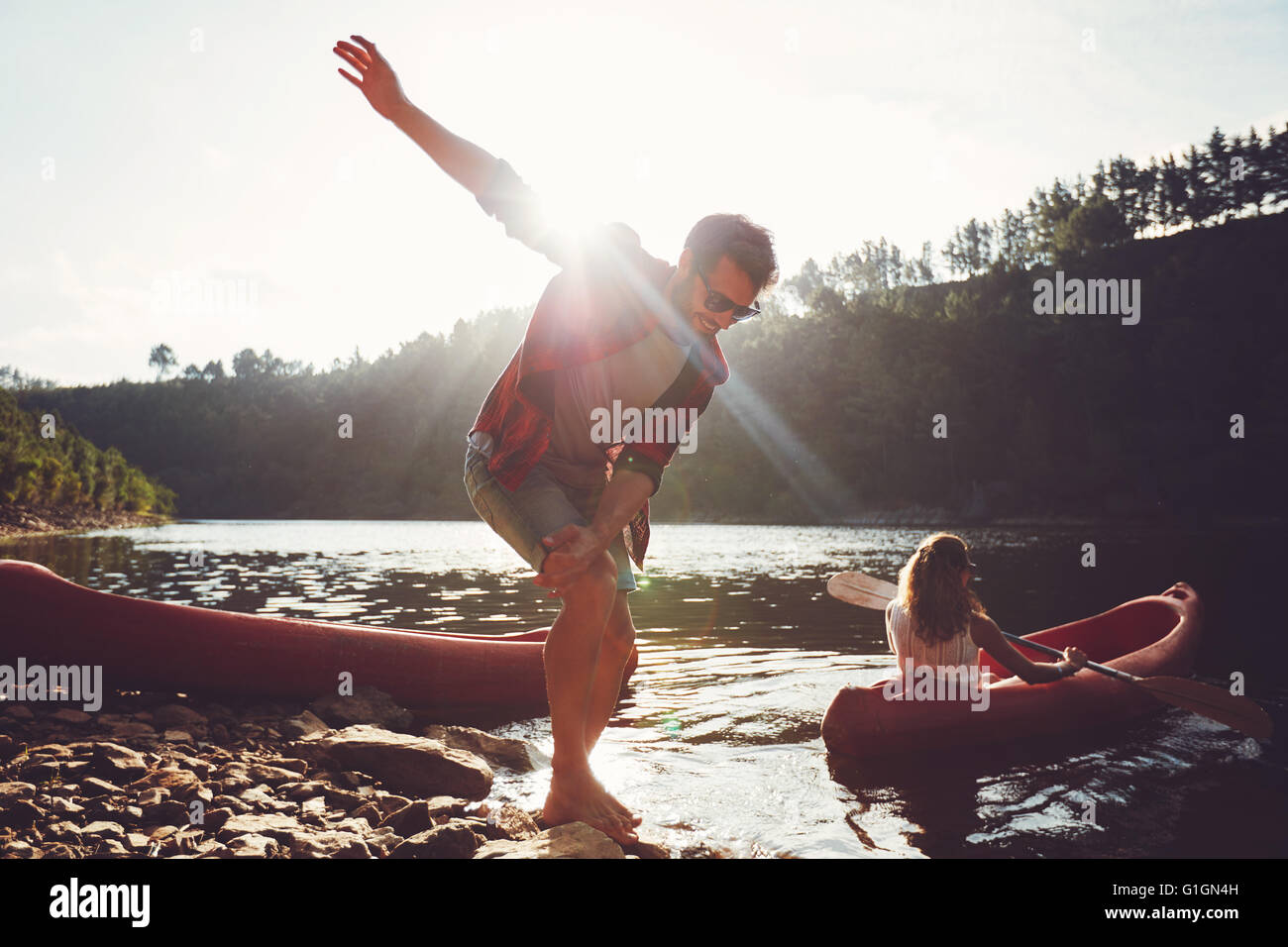 Jeune homme debout sur le bord du lac avec femme canoë. Jeune couple bénéficiant d'une journée d'été au bord du lac. Banque D'Images