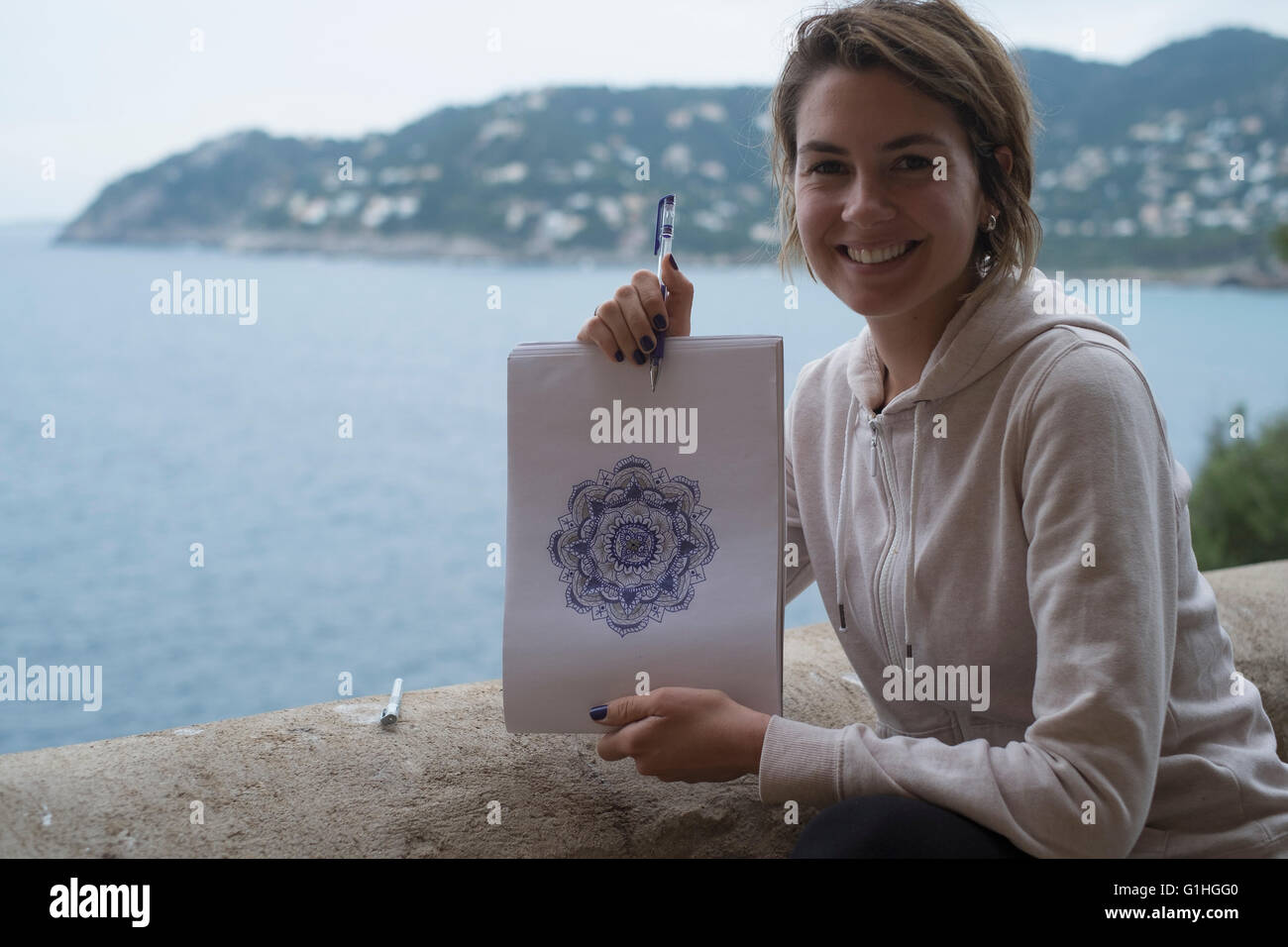 Jeune femme dessinant un mandala sur un bloc avec un crayon violet à l'extérieur, avec la mer derrière elle Banque D'Images