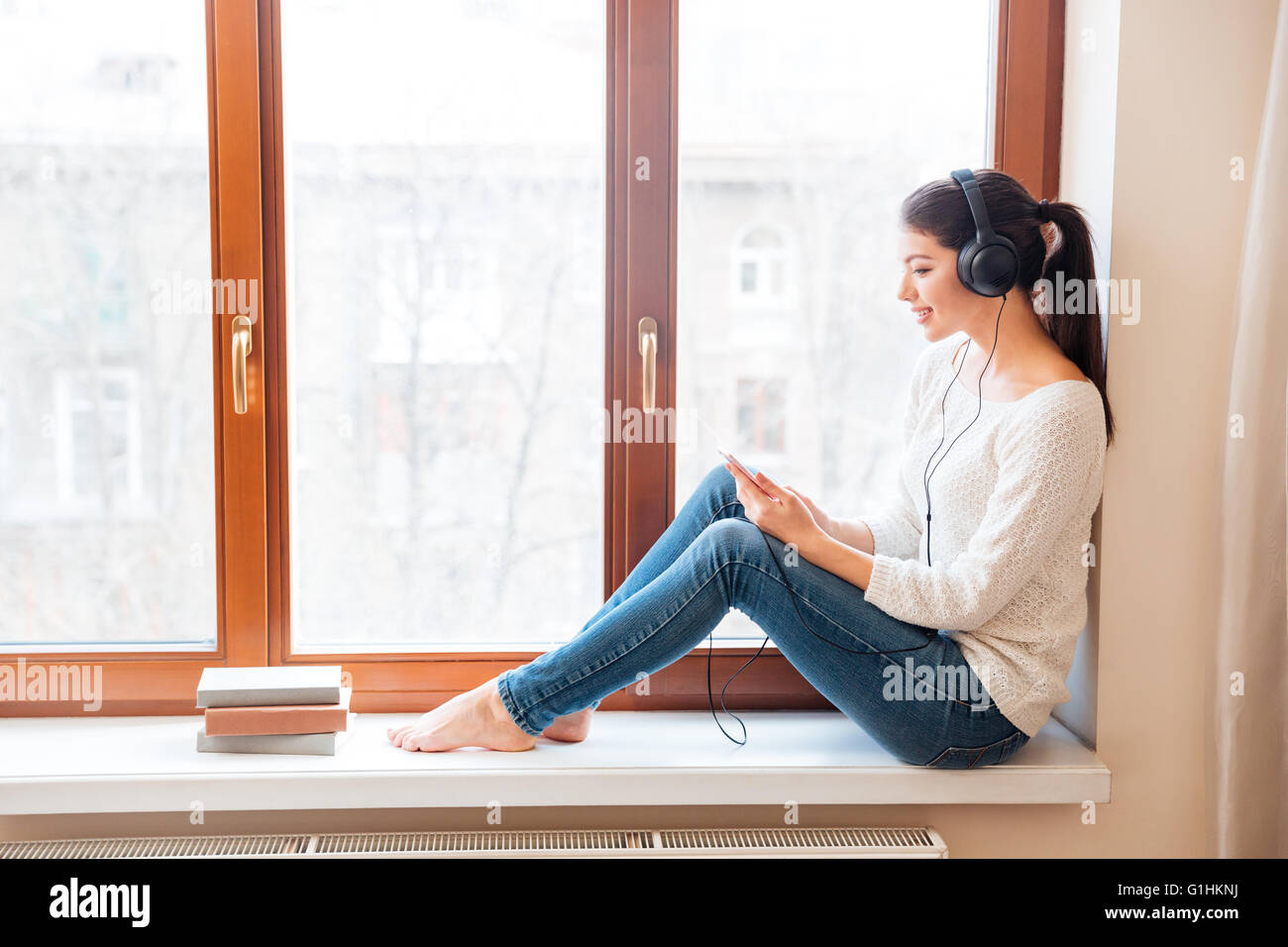 Happy woman sitting on windowsill et utilisation de smartphone avec un casque à la maison Banque D'Images