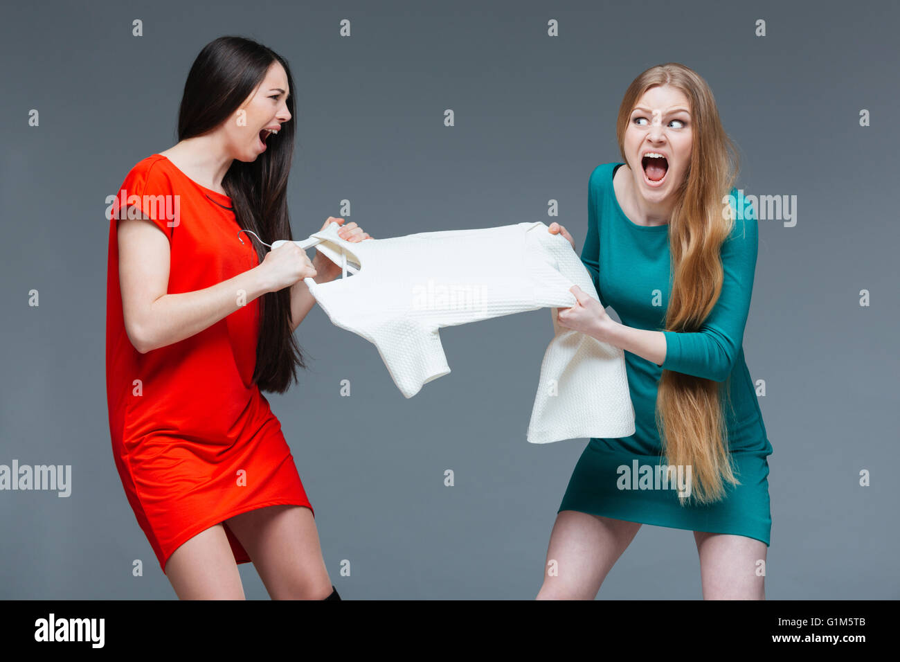 Deux jolies jeunes femmes en colère avec de longs cheveux de se quereller et de se battre pour robe blanche sur fond gris Banque D'Images