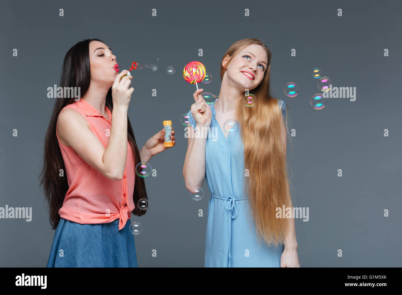 Deux belles jeunes femmes gaies colorées de l'alimentation et de sucette sur fond gris blowing bubbles Banque D'Images