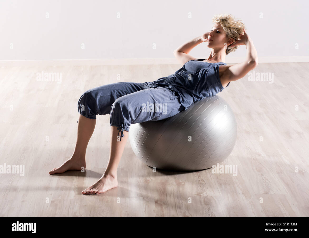 Jeune femme aux pieds nus faisant asseoir ups avec les mains derrière la tête sur grand ballon de stabilité de couleur grise sur plancher de bois franc Banque D'Images