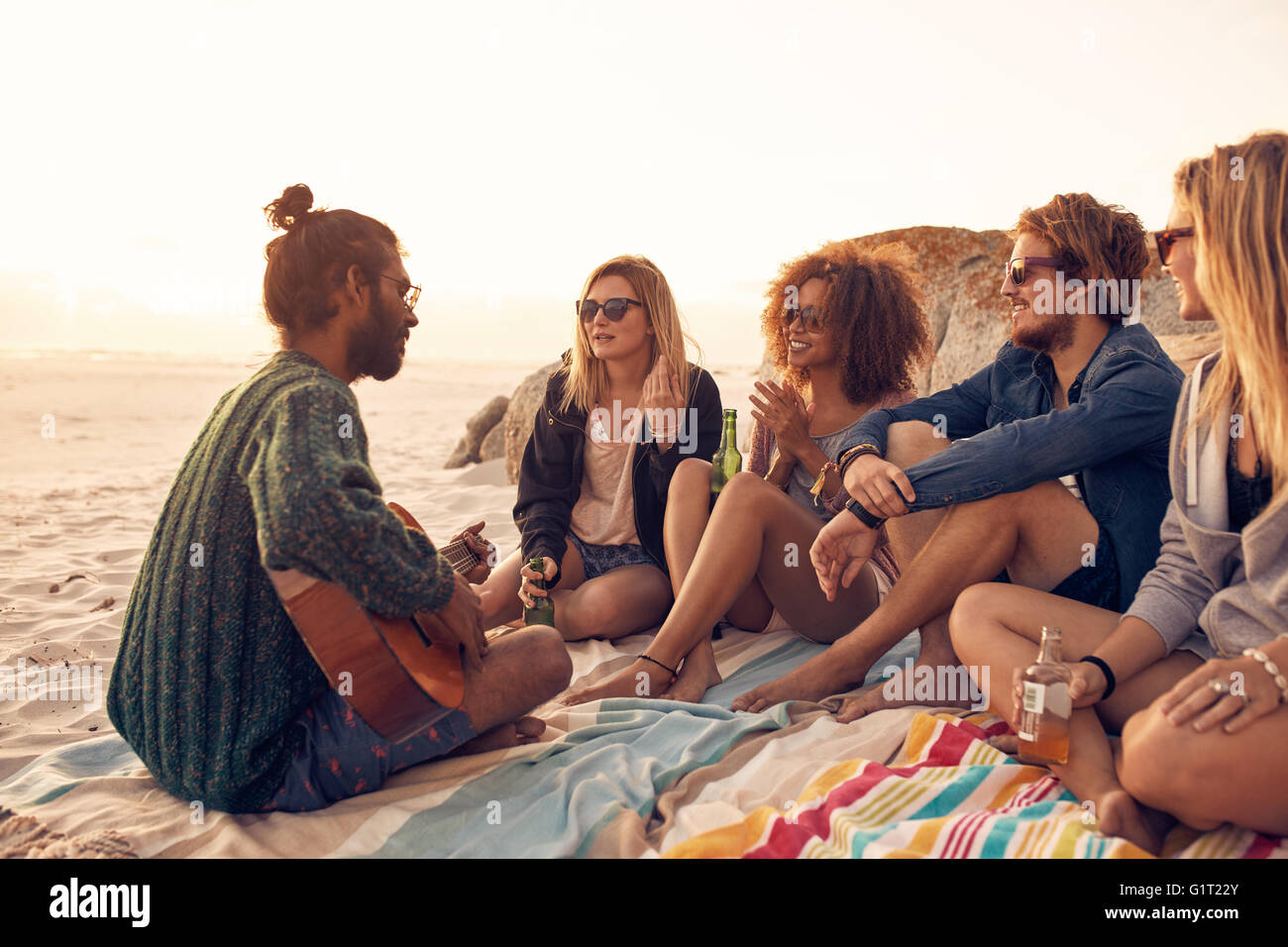 Groupe d'amis se détendre à une plage de sunset party avec jeune homme jouant de la guitare. Les jeunes ayant une partie sur la plage. Banque D'Images