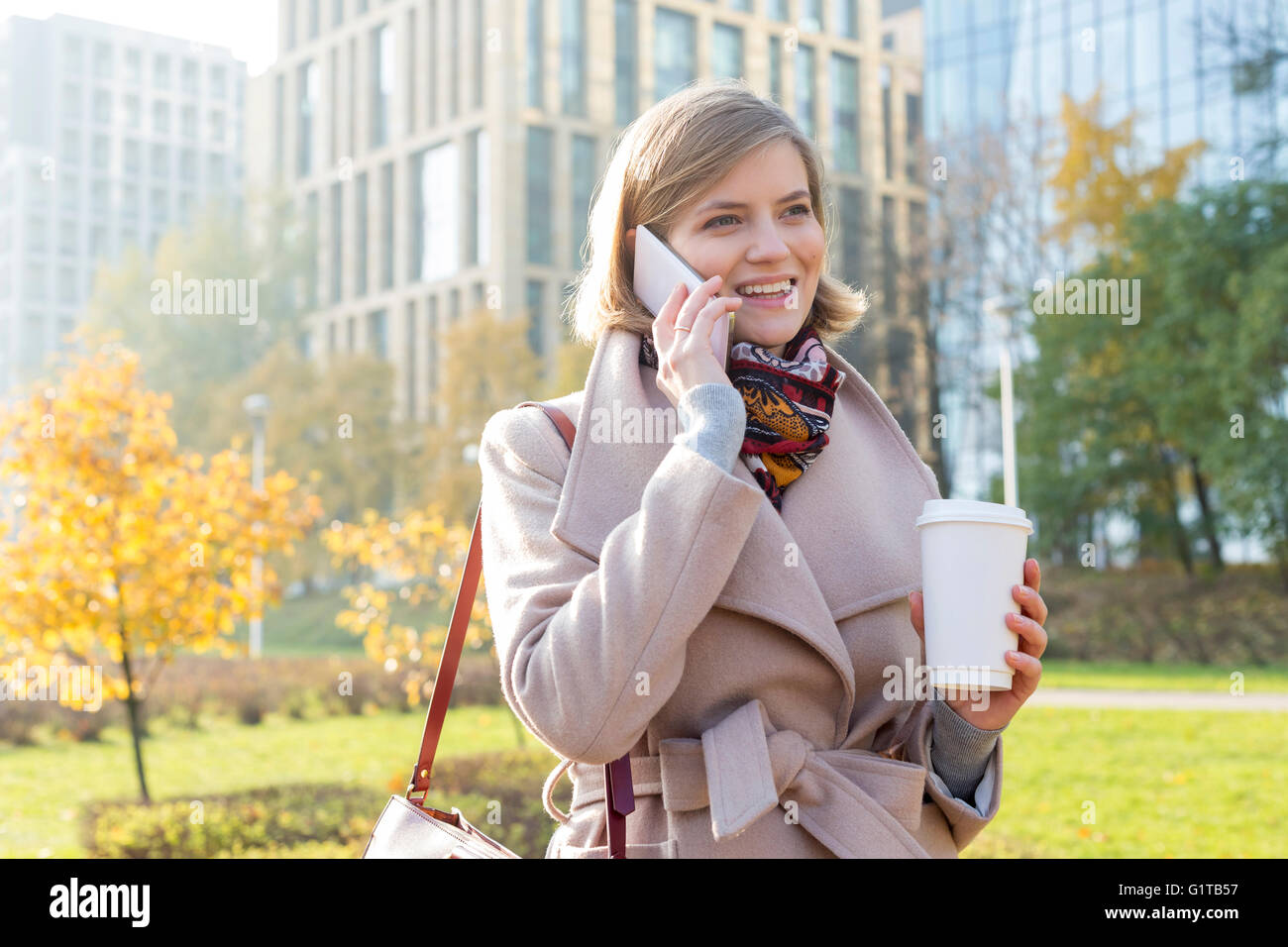 Smiling businesswoman with coffee talking on cell phone in city park Banque D'Images