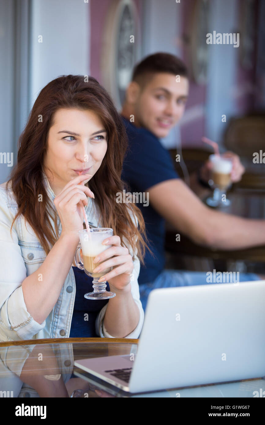 Belle jeune femme dans des vêtements décontractés assis dans street cafe avec ordinateur portable et verre, souriant au courant Banque D'Images