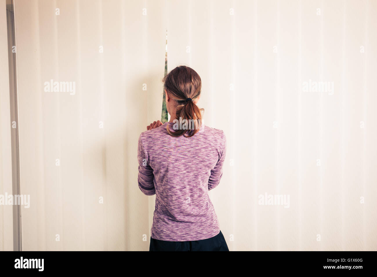 Une jeune femme se tient debout par les stores d'une fenêtre du sol au plafond Banque D'Images