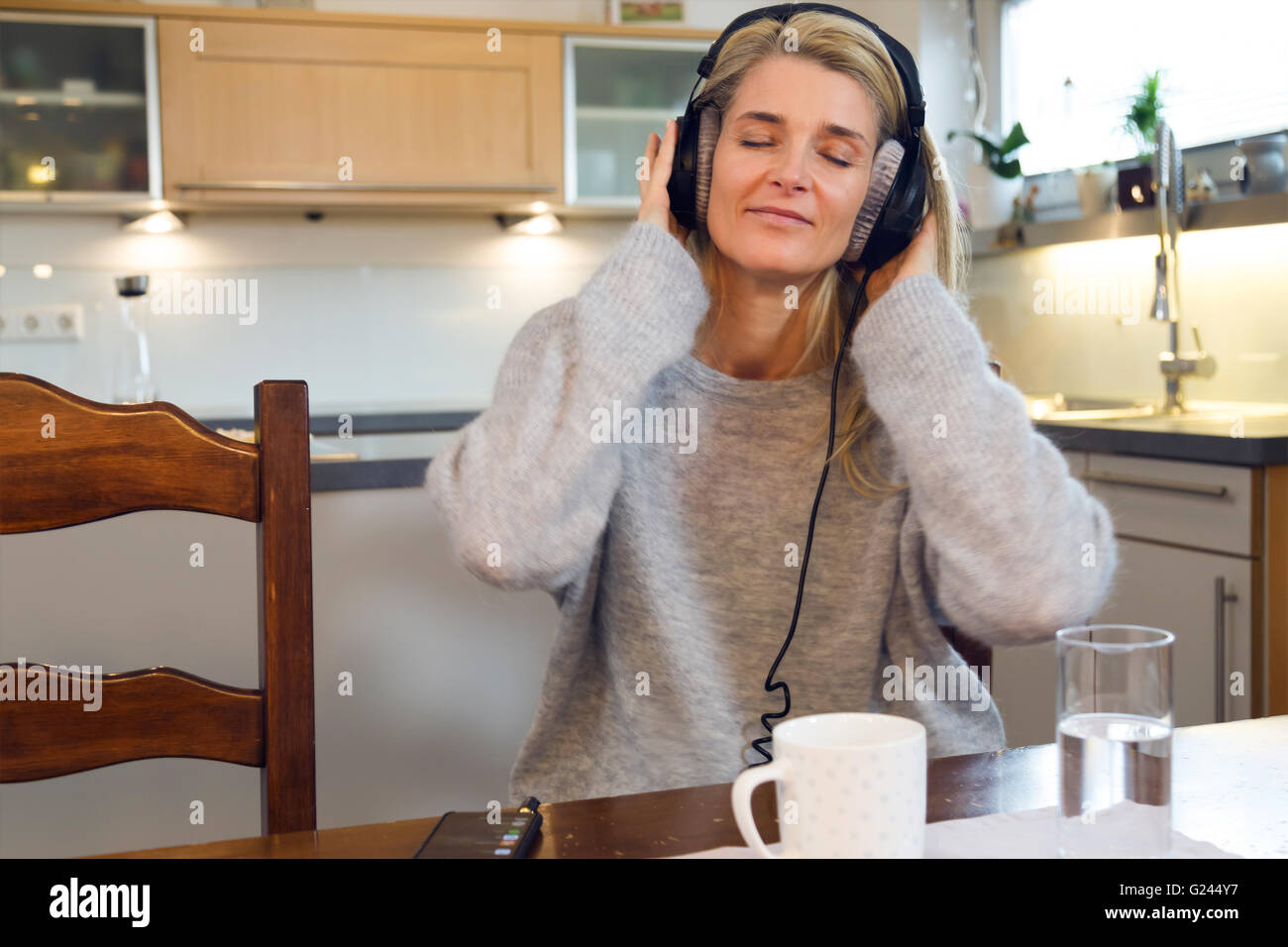 Portrait de femme d'âge moyen détendue avec des écouteurs assis à table de cuisine et de l'écoute de la musique sur votre smartphone. Tasse de café Banque D'Images