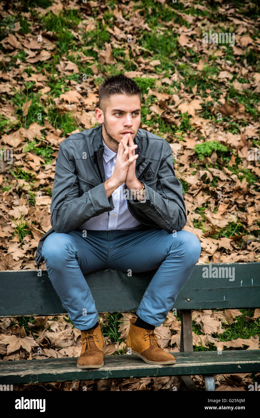 Beau jeune homme assis sur un banc dans le parc en automne, avec les feuilles mortes tout autour de Banque D'Images