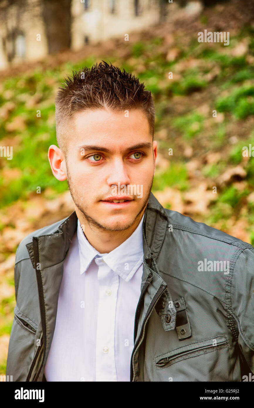 Beau jeune homme assis sur un banc dans le parc en automne, avec les feuilles mortes tout autour de Banque D'Images