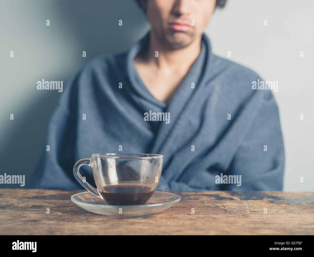 Un jeune homme fatigué portant un peignoir est assis à une table et tente de se réveiller en ayant une tasse de café Banque D'Images