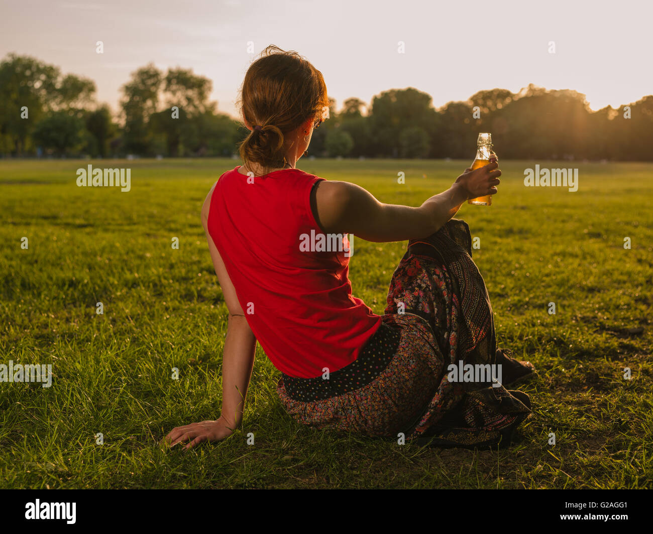 Une jeune femme est en train de boire d'une bouteille en position assise sur l'herbe dans un parc et d'admirer le coucher du soleil Banque D'Images