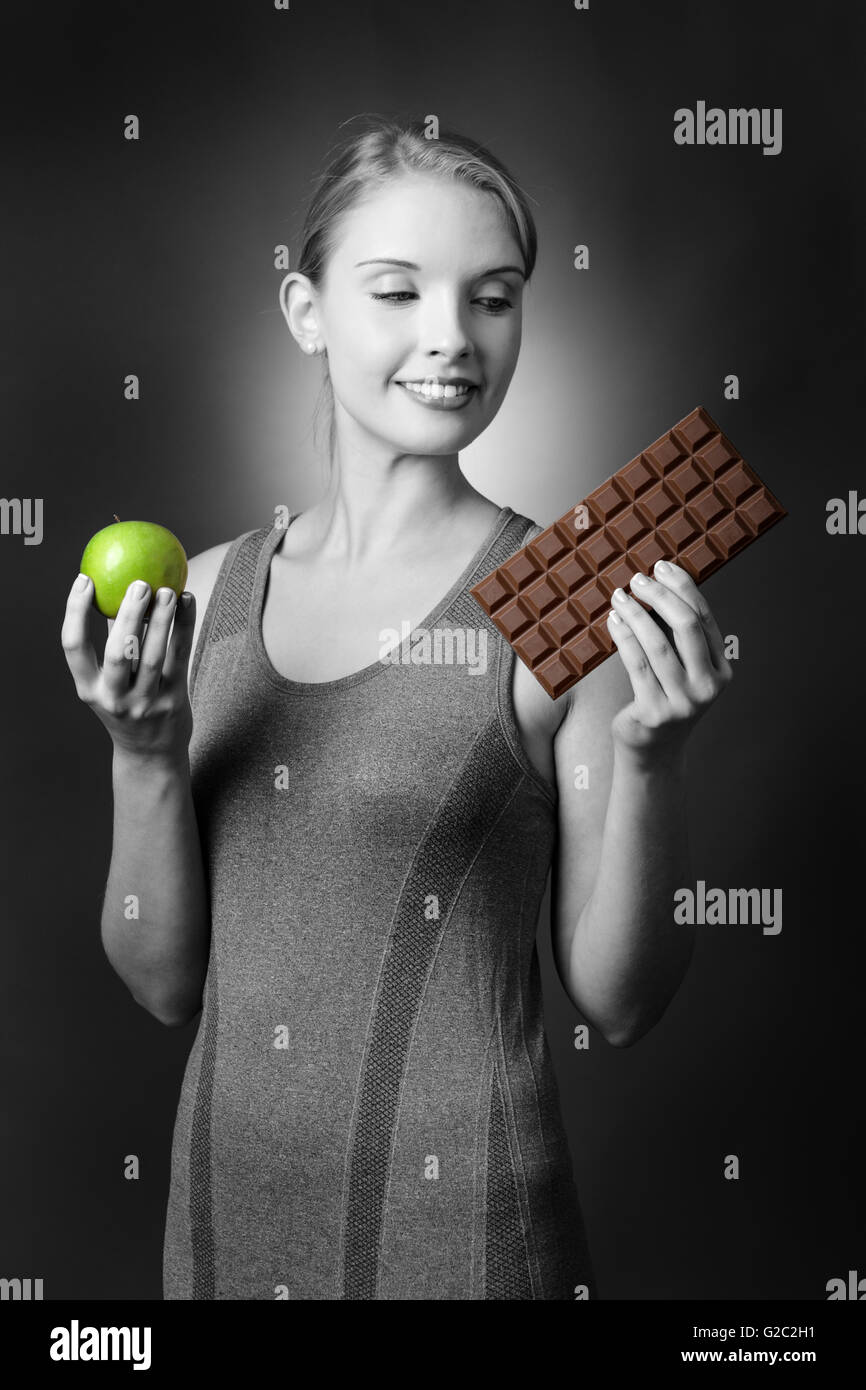 Portrait d'un joli modèle de remise en forme tenant une pomme dans une main et d'être tentés par une grande barre de chocolat dans l'autre. Banque D'Images