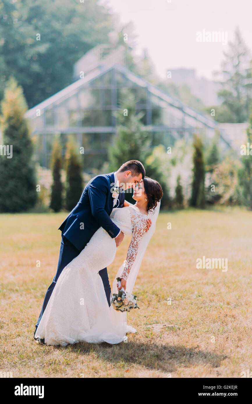 Belle Mariée avec une superbe robe de mariée blanche et le marié en costume bleu élégant posant en plein air, sur la pelouse. L'arrière-plan à effet de serre Banque D'Images