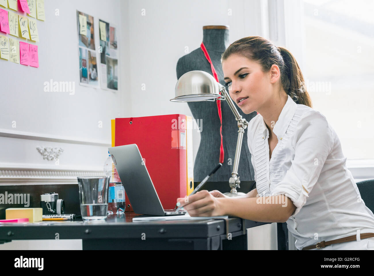 Une jeune femme d'affaires créatifs est assis à son bureau au milieu d'articles de couture lors de l'écriture dans sa note pad Banque D'Images
