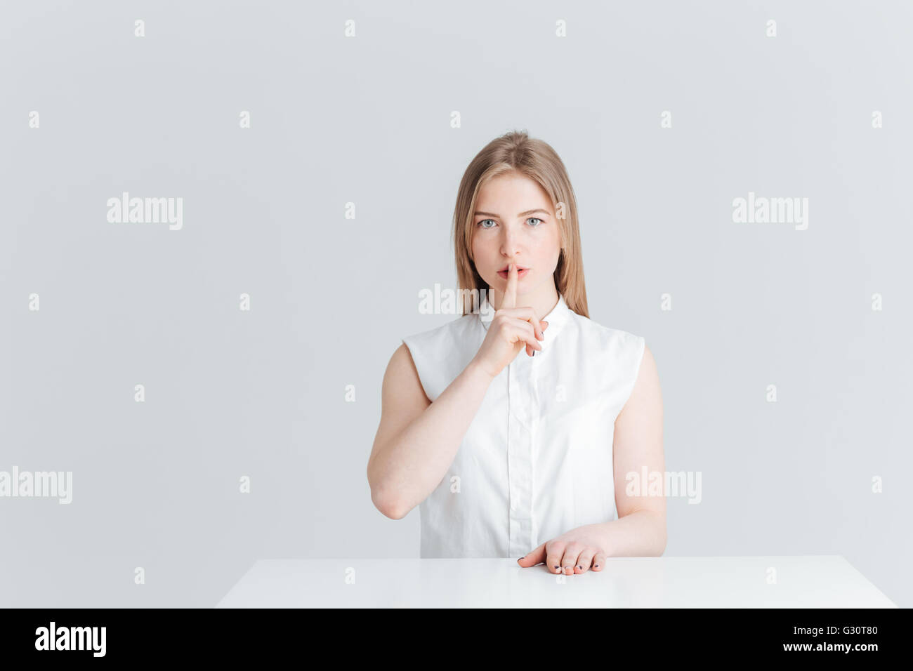 Jeune femme assise à la table et montrant doigt sur lèvres isolé sur fond blanc Banque D'Images