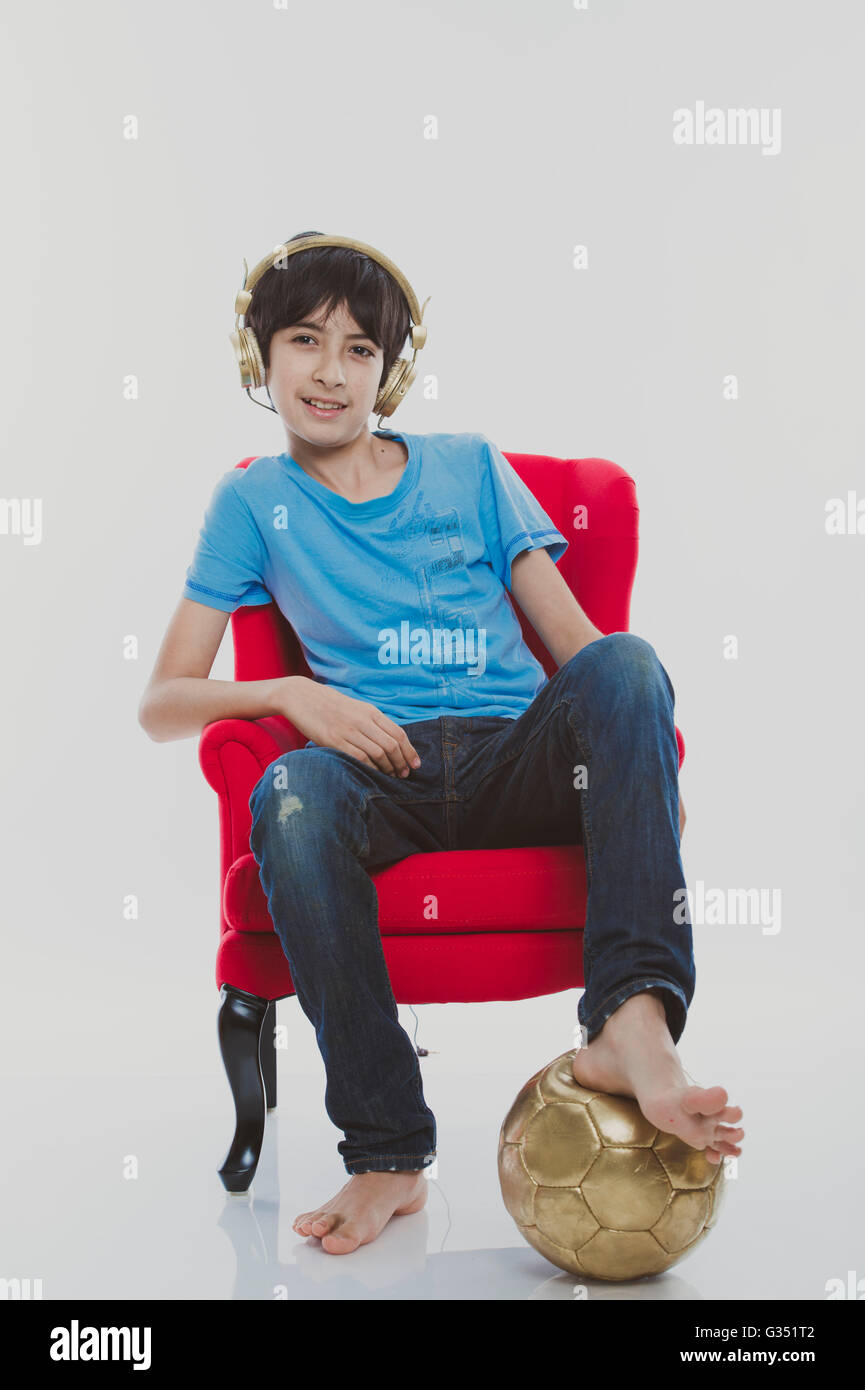 Garçon, 12 ans, assis sur une chaise rouge portant un casque d'or avec un football d'or Banque D'Images