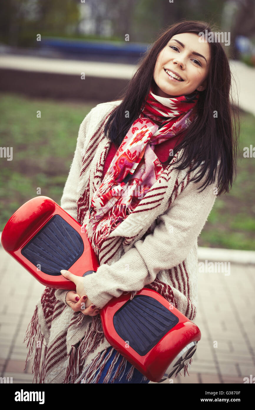 Modèle féminin moderne rouge holding electric mini hover board scooter dans les mains tout en marchant dans le parc. Populaire nouveau gadget. Girl porte des vêtements de style Boho Chic. Banque D'Images