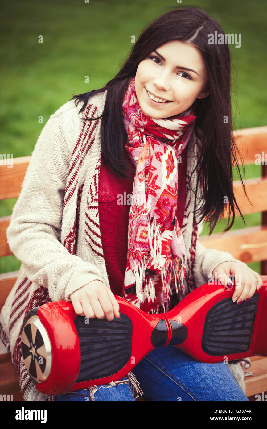 Modèle féminin tenue rouge mini électrique moderne ou segway hover board scooter dans les mains alors qu'il était assis sur un banc de parc verdoyant.Girl style Boho tendances vêtements. Banque D'Images