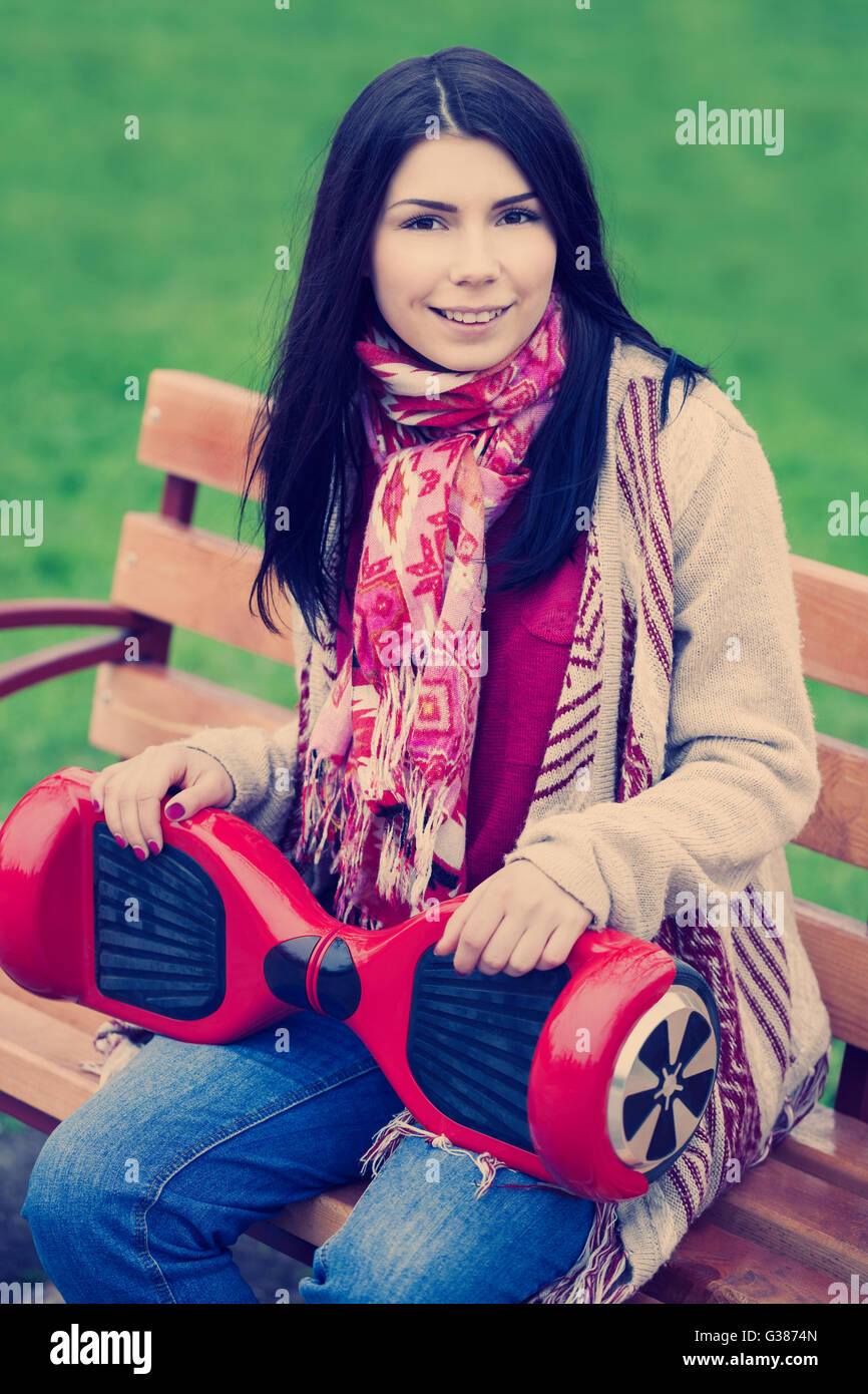 Jeune fille brune holding electric moderne rouge mini hover board scooter dans les mains tout en restant assis sur le banc de parc. Nouvelles populaires electric city transport. Fille porte des vêtements de style Boho de tendances. Banque D'Images