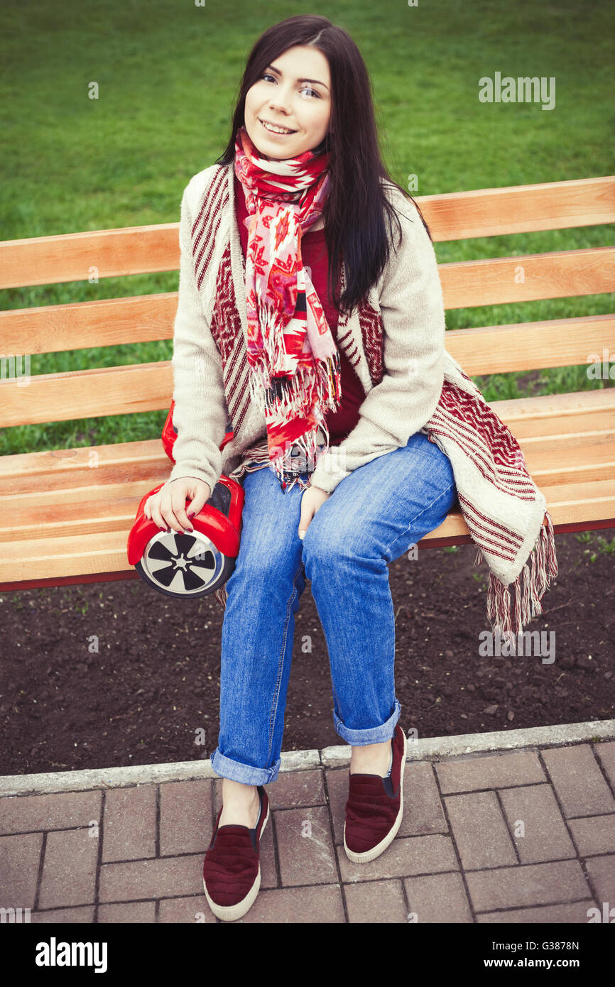 Modèle féminin moderne rouge holding electric mini hover board scooter dans mains assis sur un banc dans un parc. Nouvelles populaires city transport. Fille porte des vêtements de style Boho de tendances. Banque D'Images