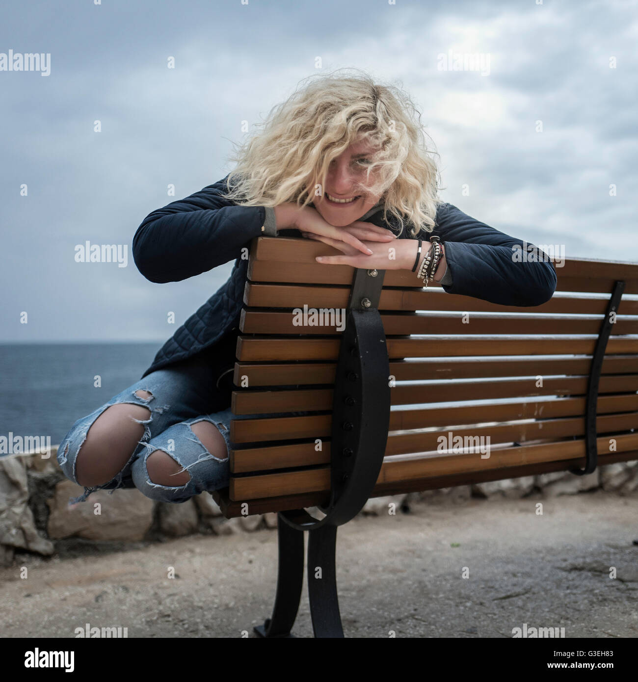 Belle fille blonde avec les cheveux bouclés en jeans déchirés est coin sur le banc près de la mer Banque D'Images