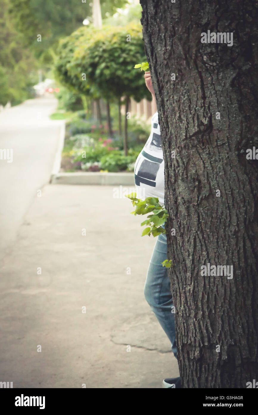 Femme enceinte à se cacher derrière l'arbre dans le parc. Concept de la grossesse. Banque D'Images