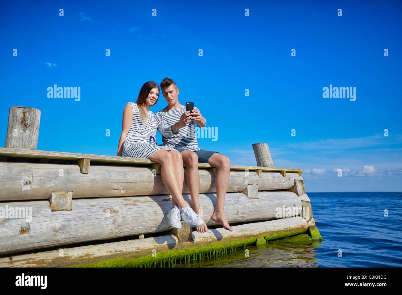 Couple sitting on wooden pier, taking self portrait using smartphone Banque D'Images