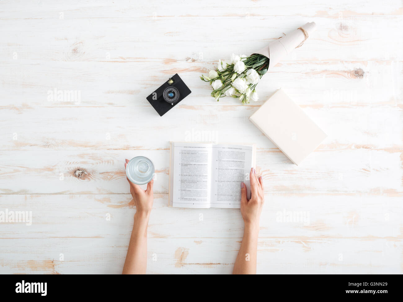 Les mains des femmes plus de tourner les pages d'un livre avec un verre d'eau, des fleurs et de la caméra sur le bureau en bois Banque D'Images