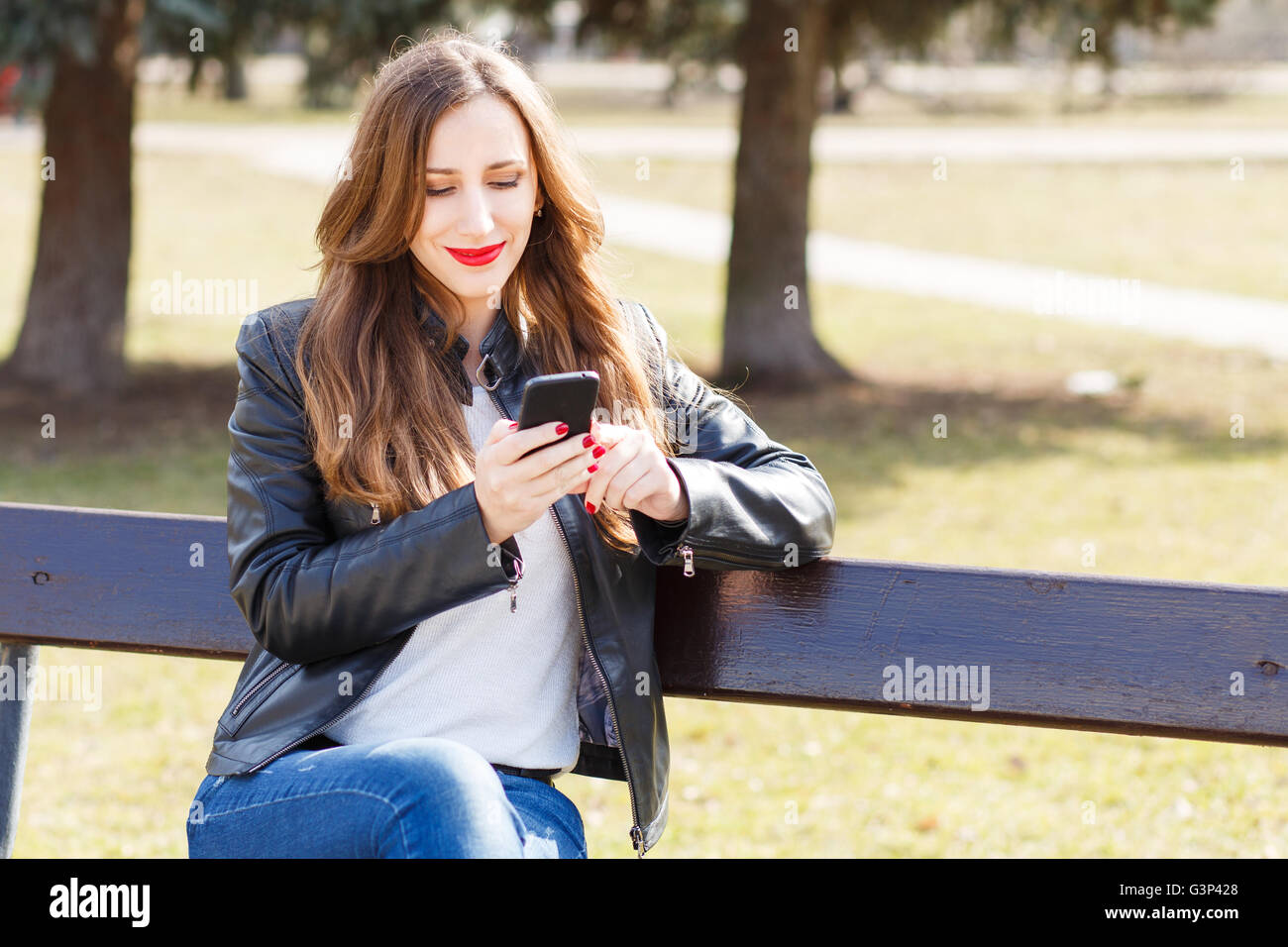 Young smiling woman using smartphone assis sur un banc de parc. Belle fille européenne texting on phone Banque D'Images
