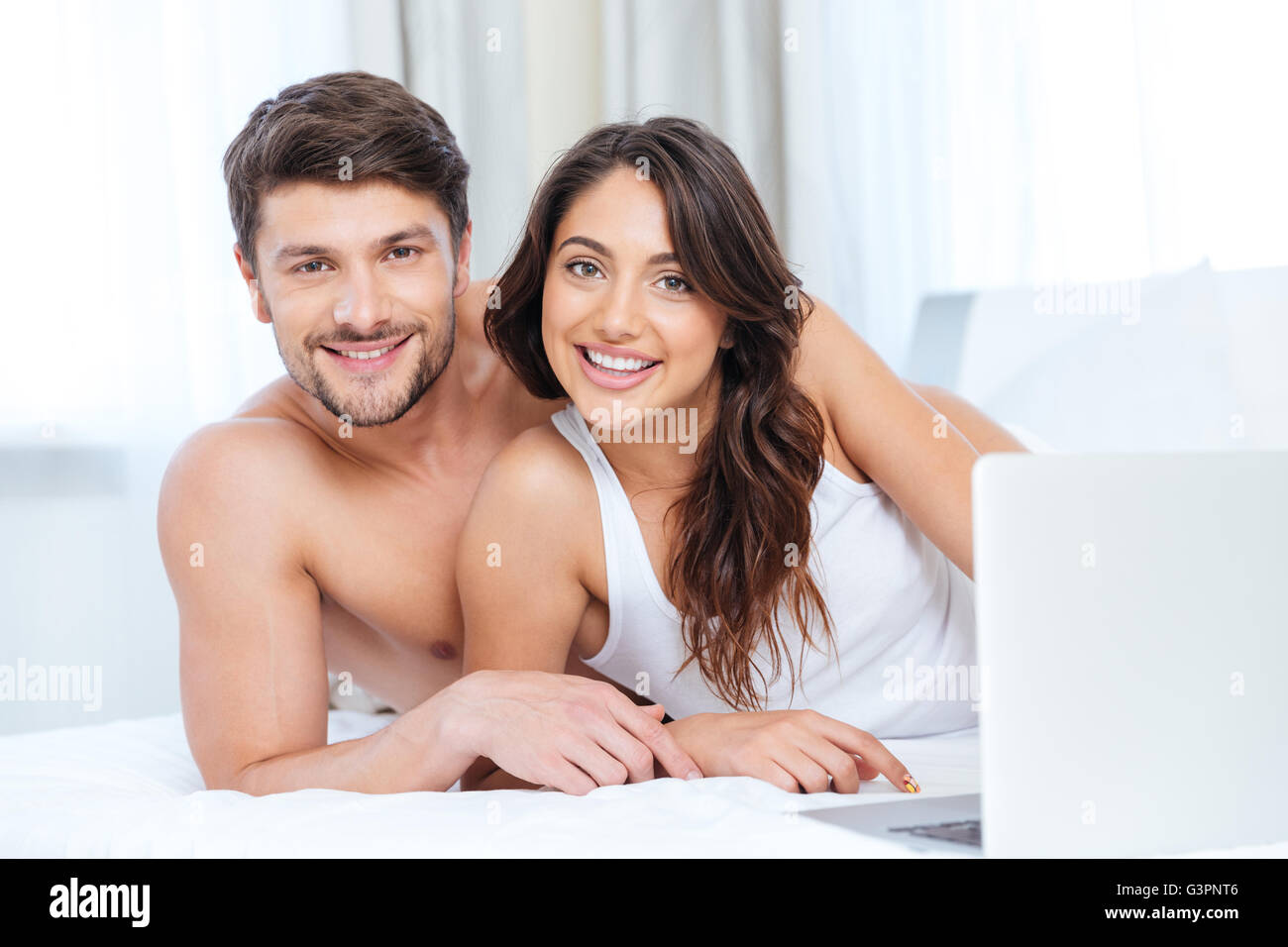 Smiling beautiful couple using laptop and looking at camera au lit à la maison Banque D'Images