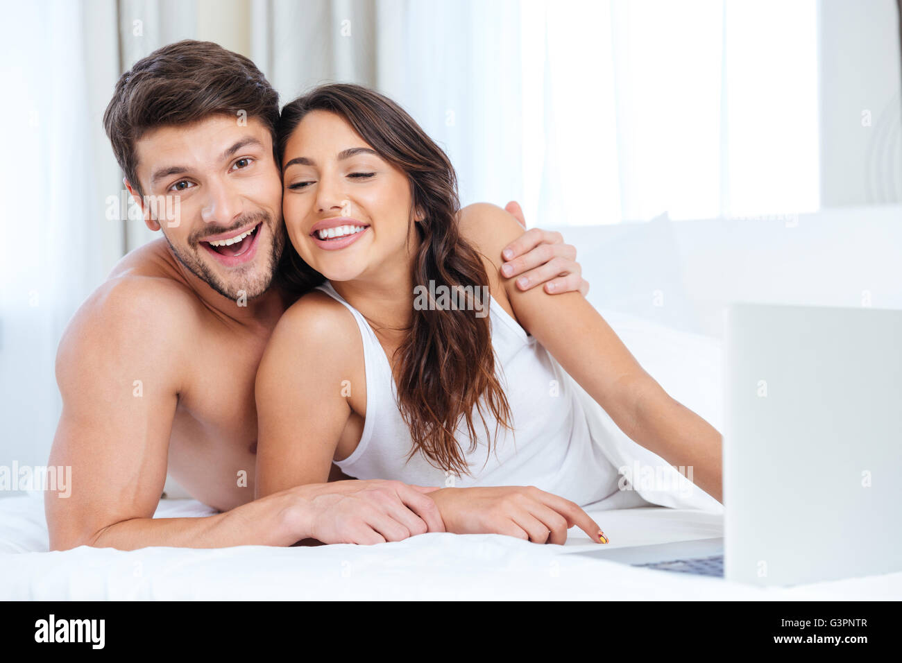 Smiling beautiful couple using laptop and looking at camera au lit à la maison Banque D'Images