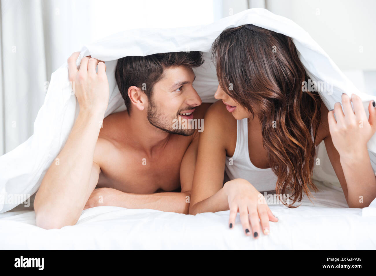 Couple heureux au lit recouvert d'une couverture sur la tête et de parler à la maison plus de lumières fond Banque D'Images