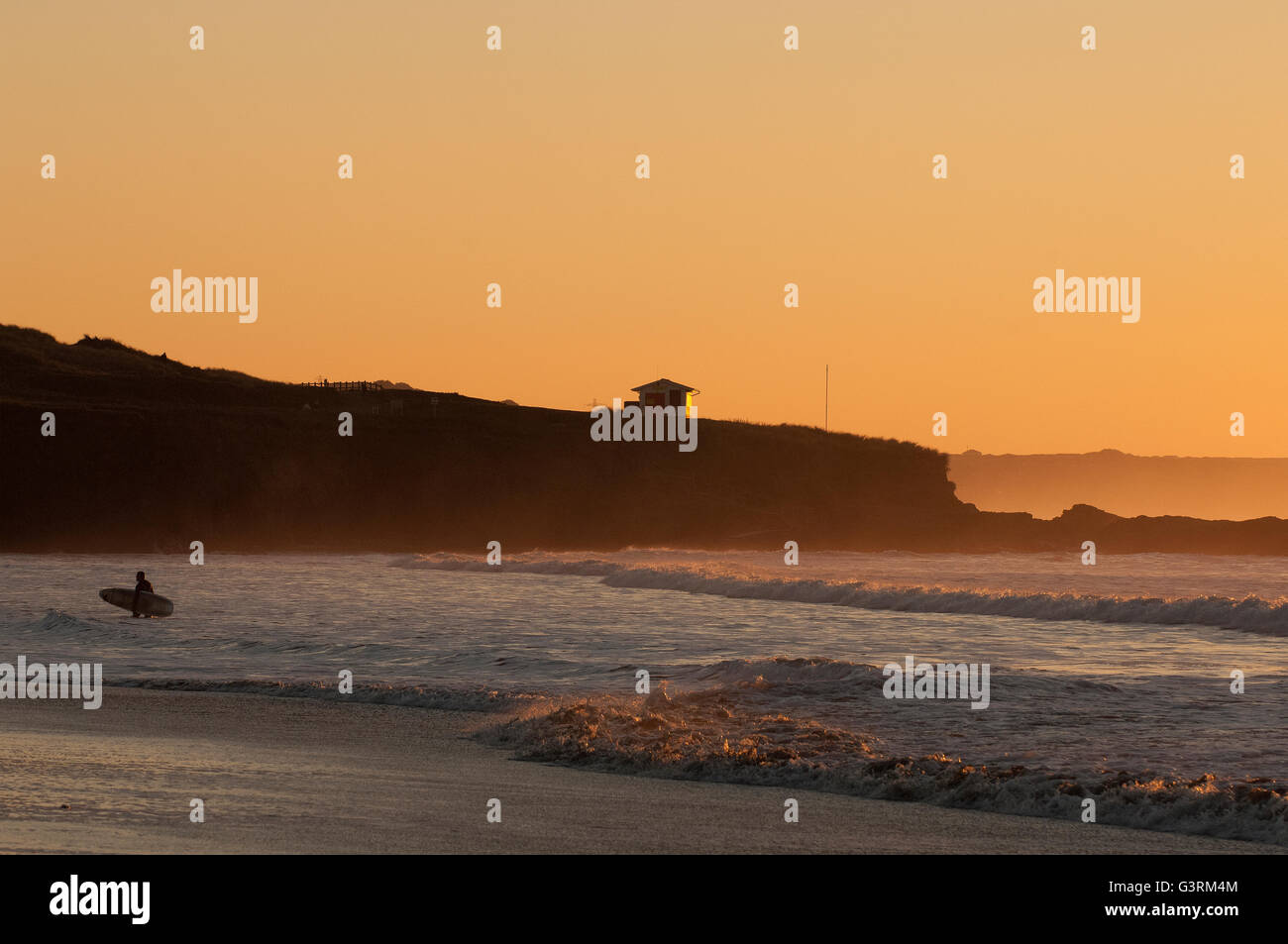 Surfer en silhouette de quitter l'eau avec le RNLI lifeguard hut baigné de soleil sur la pointe à Gwithian plage au coucher du soleil Banque D'Images