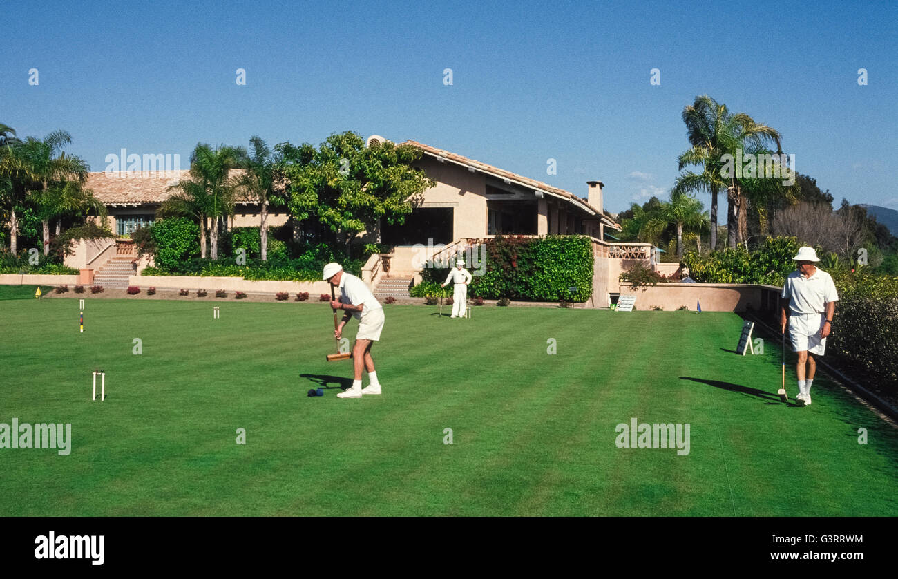 La traditionnelle partie de croquet est une activité populaire pour les membres de la section locale du club de croquet qui s'habillent en blanc tous les vêtements, ainsi que pour les clients du Rancho Valencia Resort & Spa, qui tous deux jouent sur le vaste terrain de croquet de cet hôtel de luxe dans le comté de San Diego, Californie, USA. Membres de l'association suivre les règles officielles de l'Association américaine pour l'Américain Six-Wicket Croquet Croquet que spécifier l'équipement à utiliser, y compris les maillets, ballons, guichets et jeu. Ici un mâle joueur club se prépare à frapper sa balle bleue avec un maillet en bois carrés à conduire une boule noire. Banque D'Images