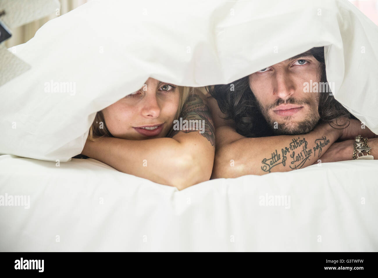 Un jeune couple cool tatoués, à moins de une couette sur un lit. Banque D'Images
