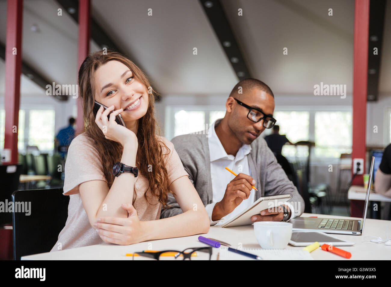 Cheerful beautiful young business woman talking on cell phone et travailler avec african man in office Banque D'Images