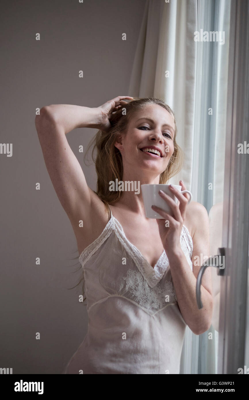 Jolie femme de boire du café par la fenêtre dans sa robe de nuit Banque D'Images
