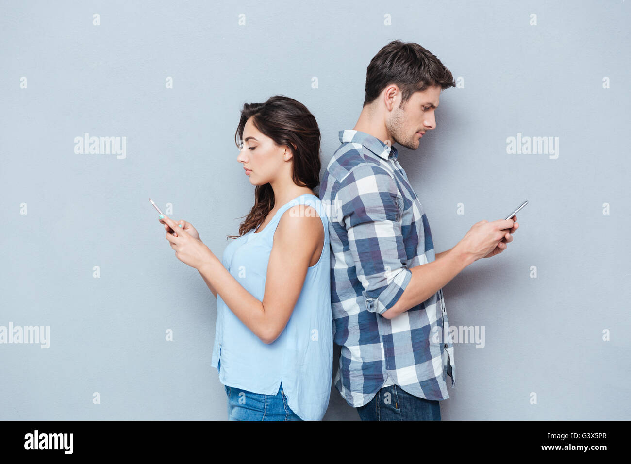 Portrait of young couple debout dos à l'autre à la recherche et le téléphone isolé sur fond gris Banque D'Images