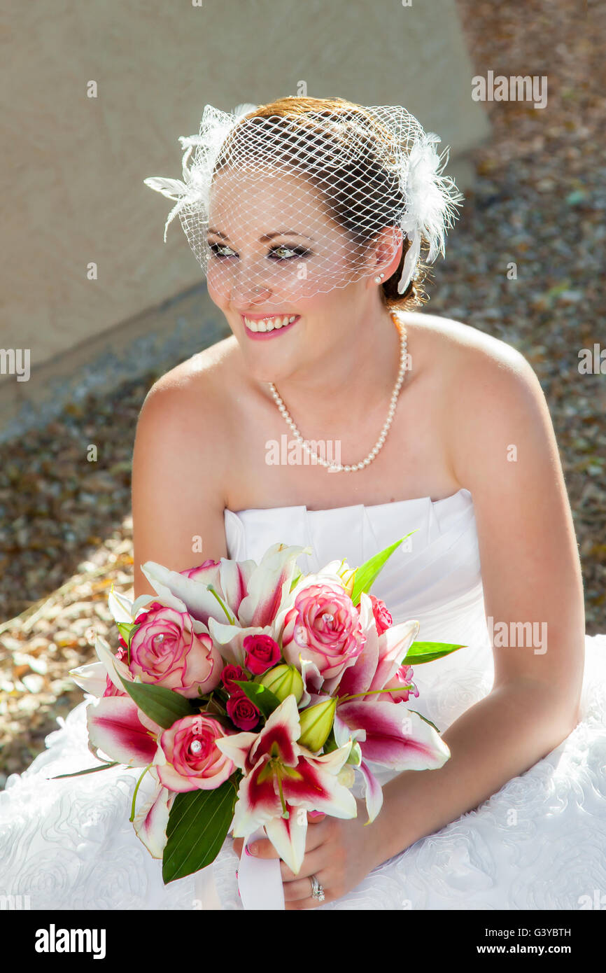 Une nouvelle mariée mariés s'accroupit pour prendre une courte pause de douloureuses chaussures. Elle a un sourire sur son visage et un beau bouquet Banque D'Images